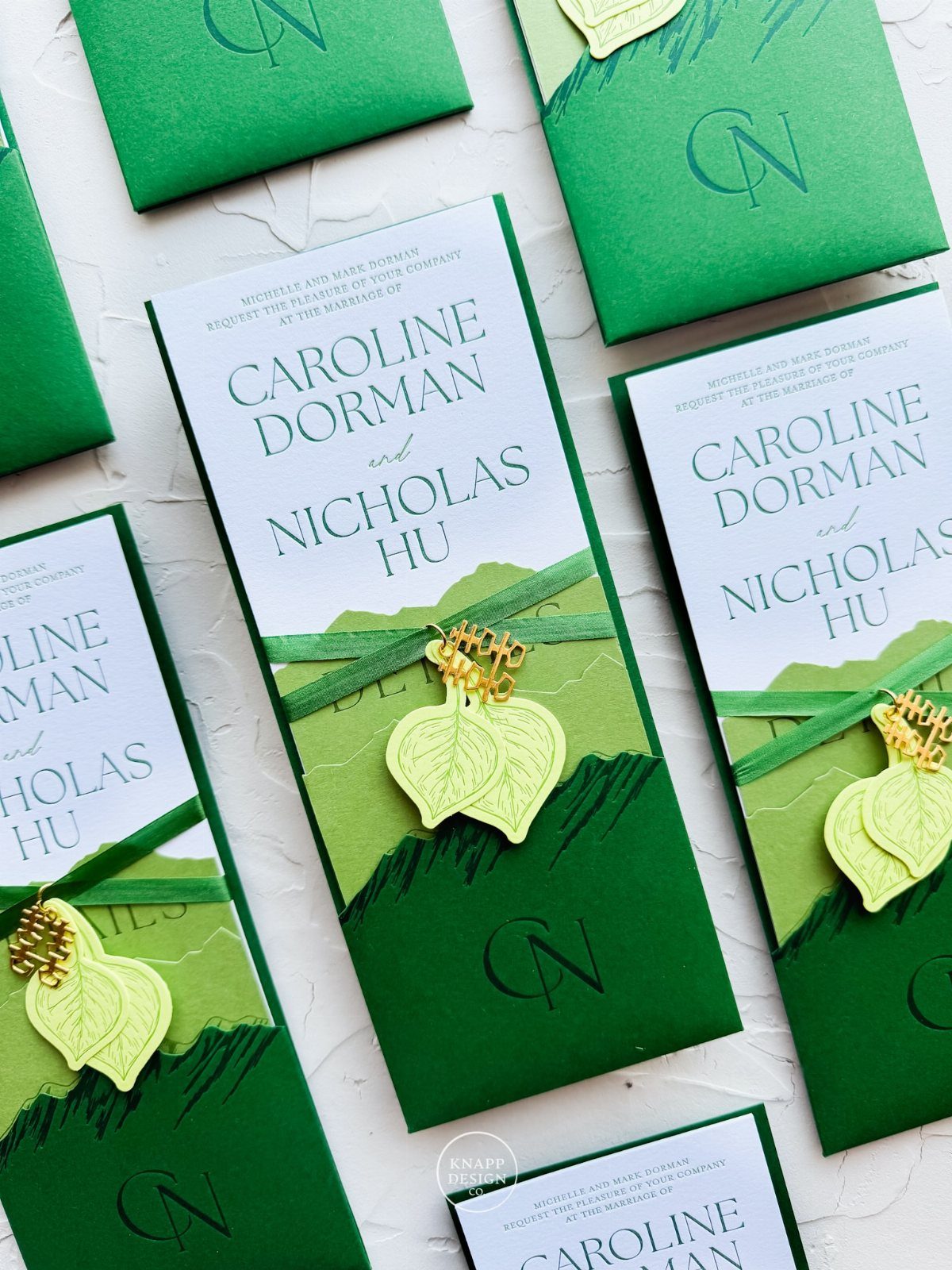 Green and white wedding invitations with teal text and leaf-shaped charms on ribbons.