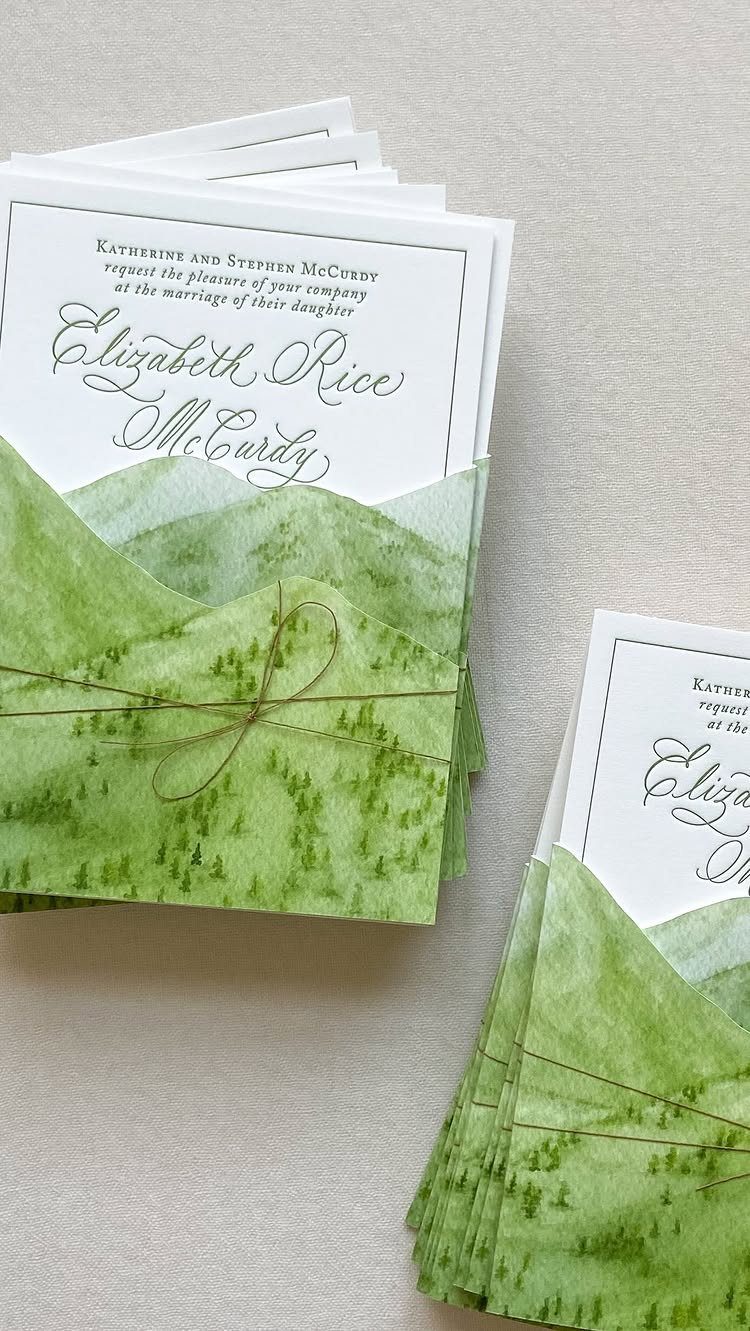 Wedding invitations with cursive text in green watercolor mountain-shaped envelopes tied with twine.