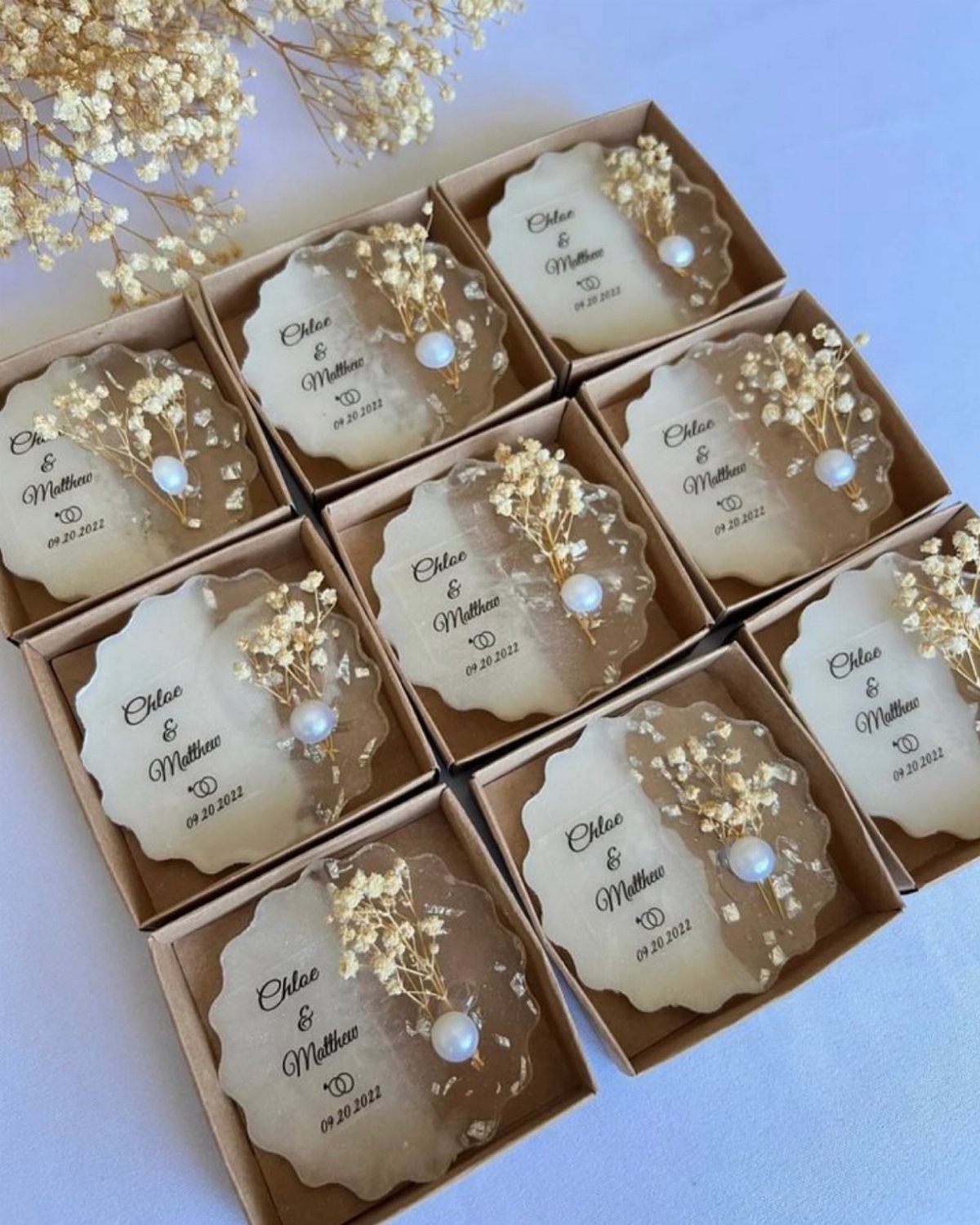 Twelve personalized resin wedding favor ornaments with dried flowers in brown gift boxes.