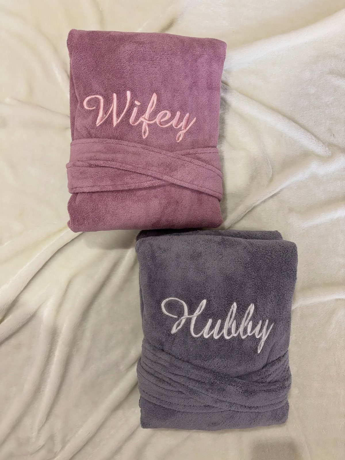 Two folded plush bathrobes embroidered with Wifey and Hubby on white fabric.