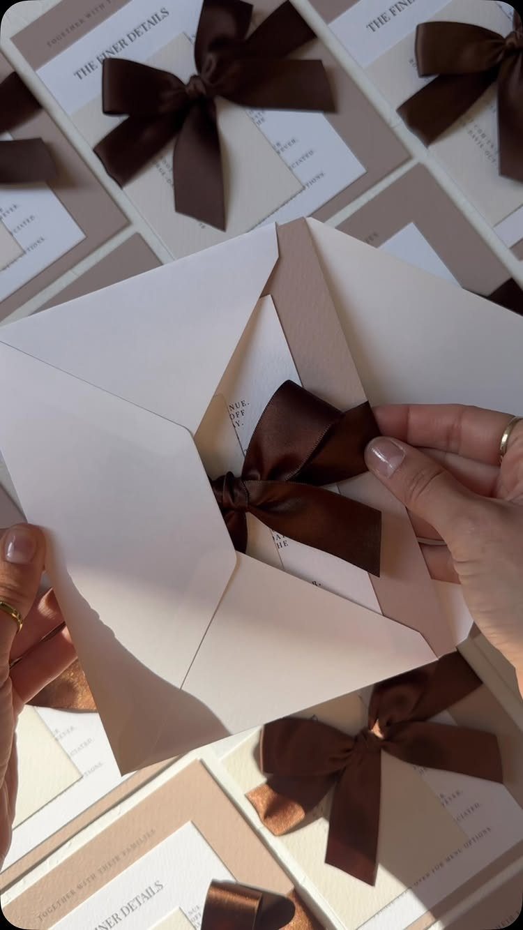 Cream wedding invitations tied with brown satin ribbons held by a person's hands.