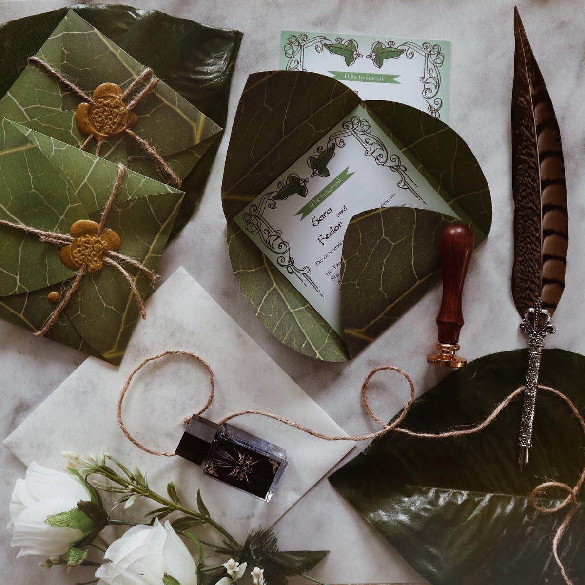 Leaf-shaped wedding invitations with gold wax seals, a quill, and white roses.