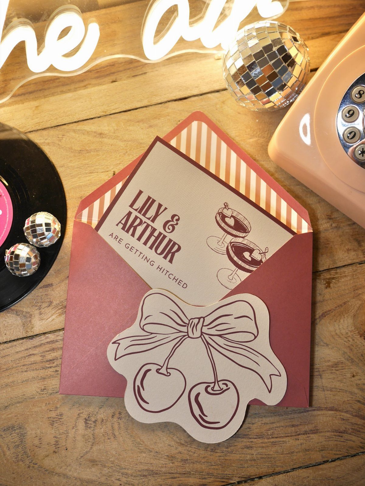 Retro wedding invitation with cocktail art in a striped pink envelope on wood.