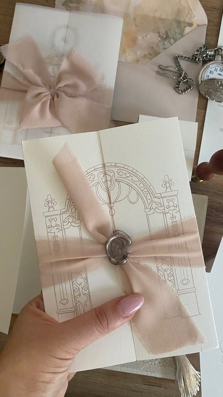 Cream wedding invitation with ornate line art, blush ribbon, and silver wax seal.