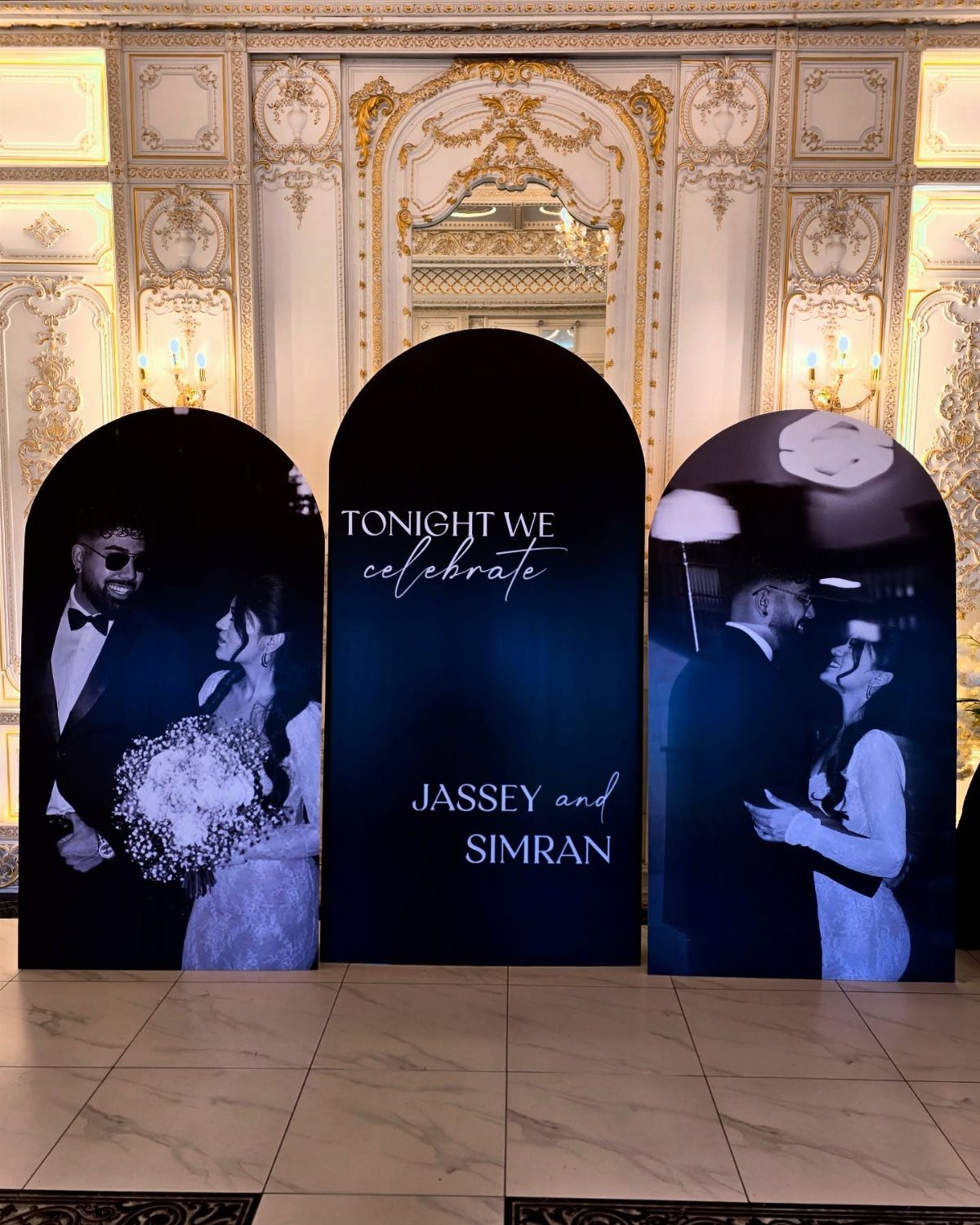 Three black arched wedding signs with photos and names in an ornate hall.