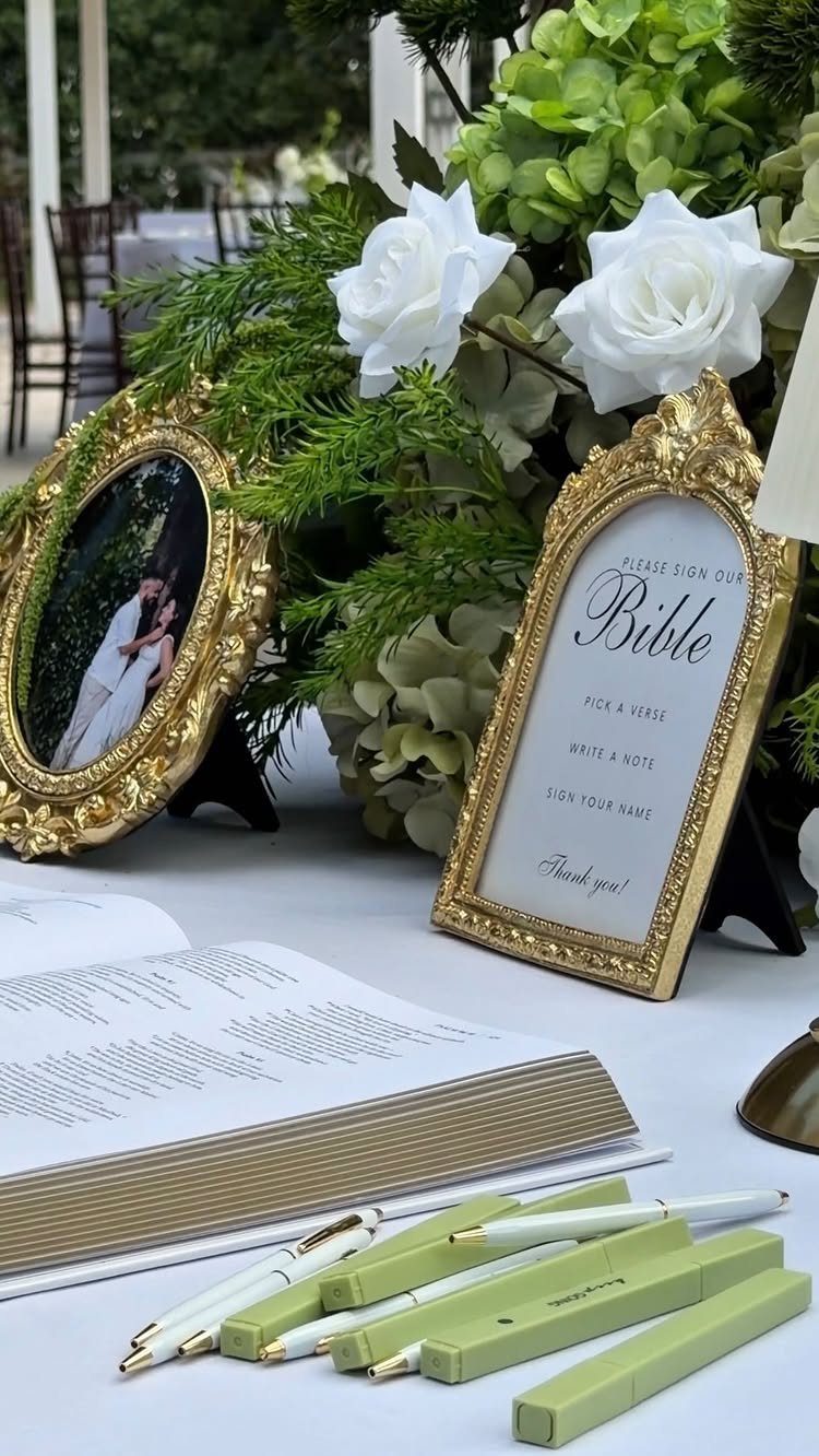 Gold framed guestbook sign next to an open bible and green pens with flowers.