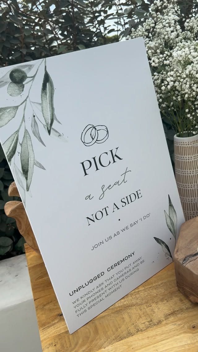 White wedding sign with greenery requesting an unplugged ceremony and open seating.