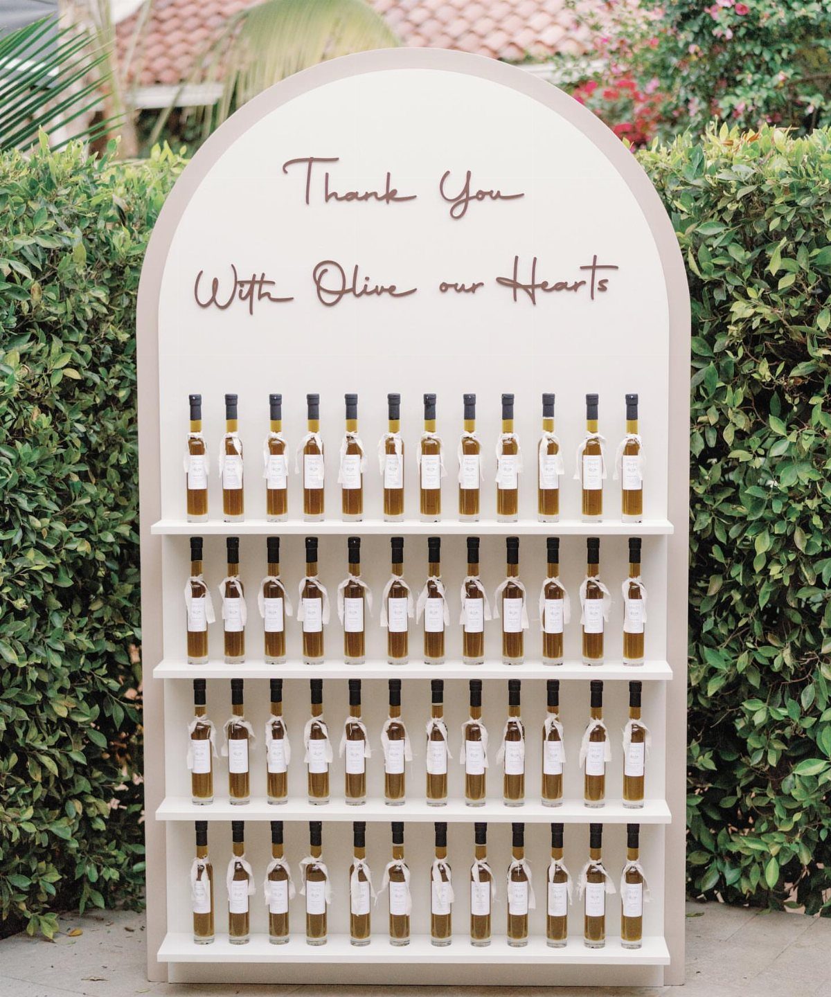 Mini olive oil bottles with white labels displayed on a wedding favor shelf.
