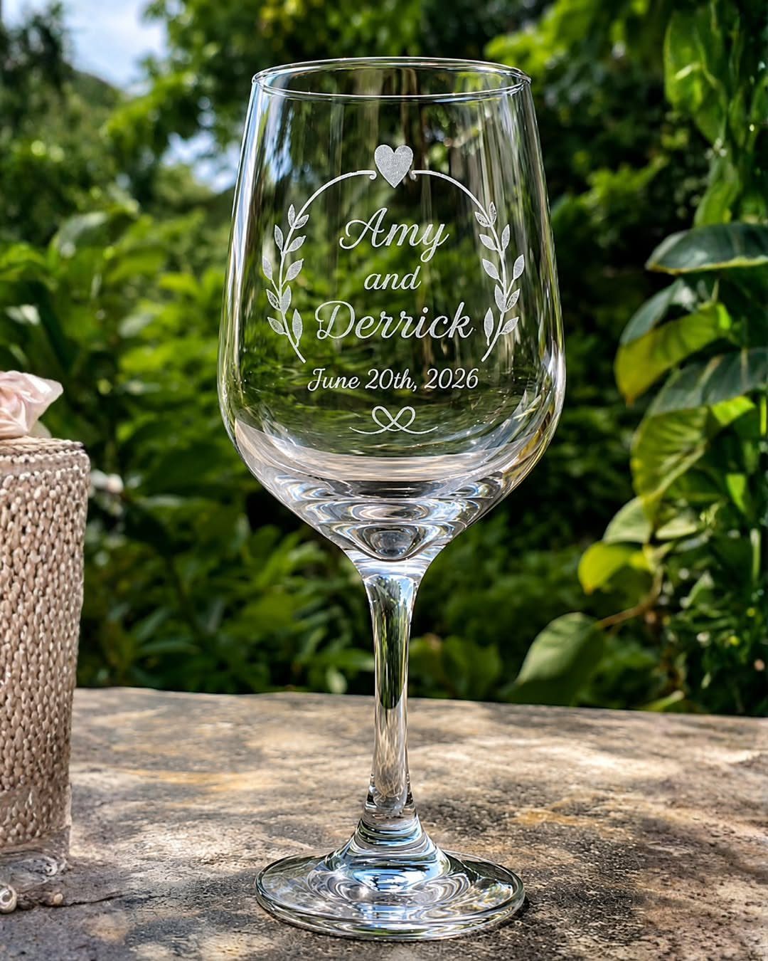 Etched wine glass with wedding names and date set against a green garden.