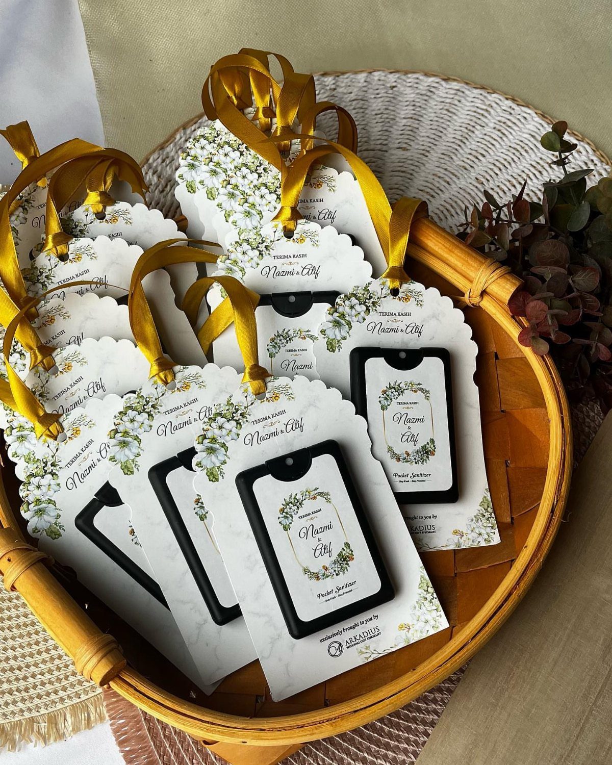 Hand sanitizer bottles with personalized floral tags and gold ribbons in a wicker basket.