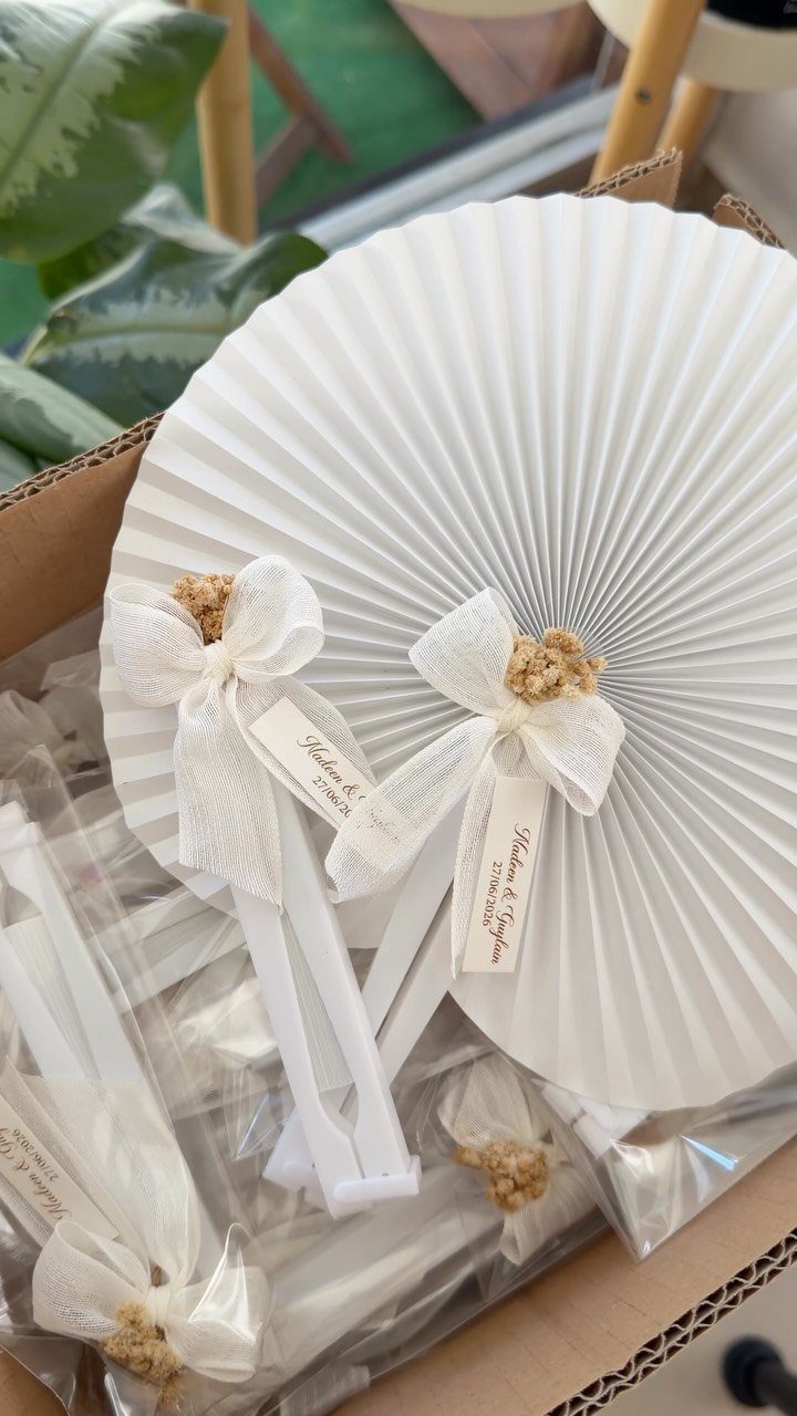 White pleated hand fans with ribbons and personalized tags in a cardboard box.