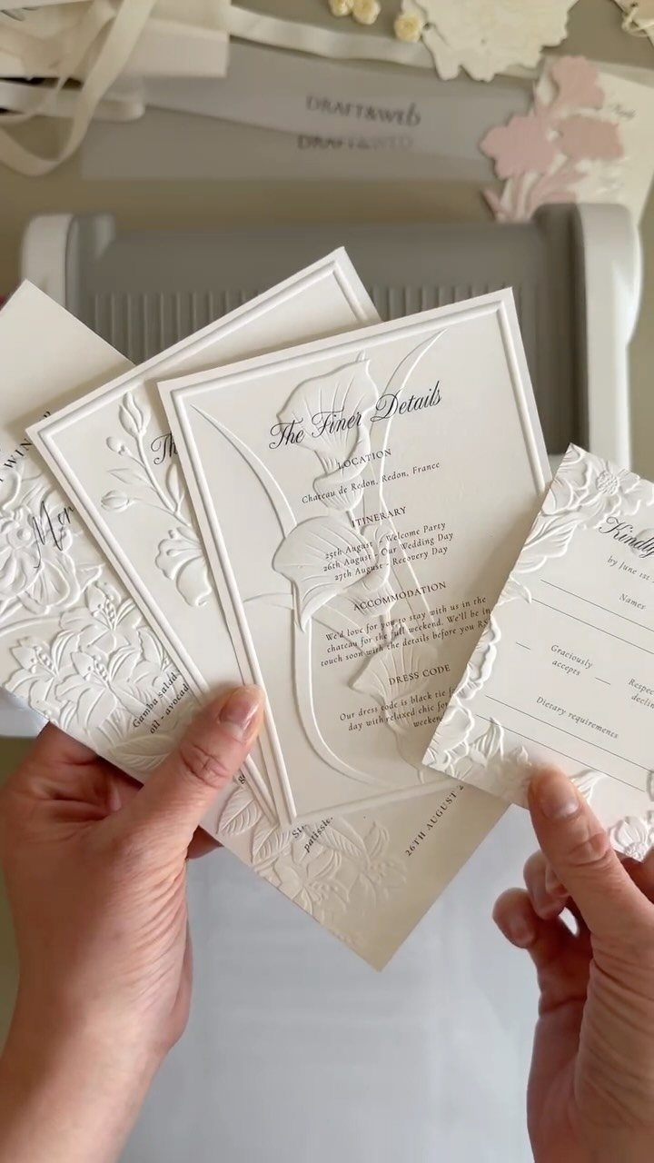 White embossed wedding invitation cards with raised floral designs held by two hands.