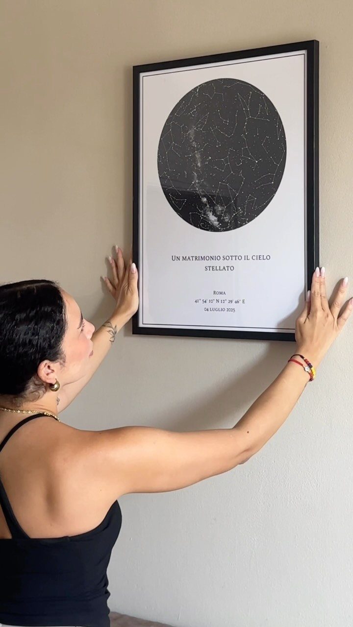 Woman hanging a framed star map with constellations on a beige wall.