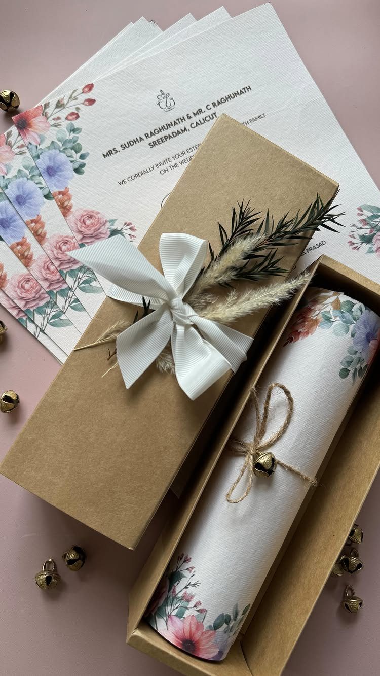 Rolled floral wedding invitation tied with twine and a gold bell in a box.