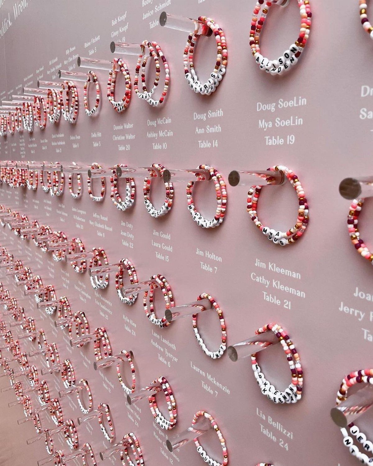 Beaded friendship bracelets displayed on a pink wall with guest names and table numbers.