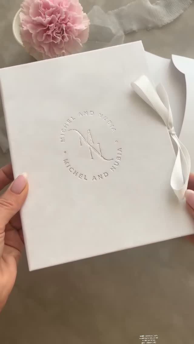 White embossed wedding invitation with a ribbon bow held by two hands.