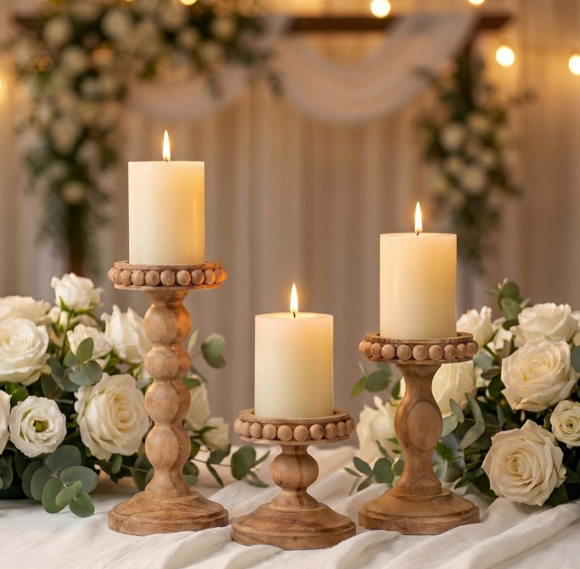 Three wooden candle holders with lit white candles surrounded by white roses.