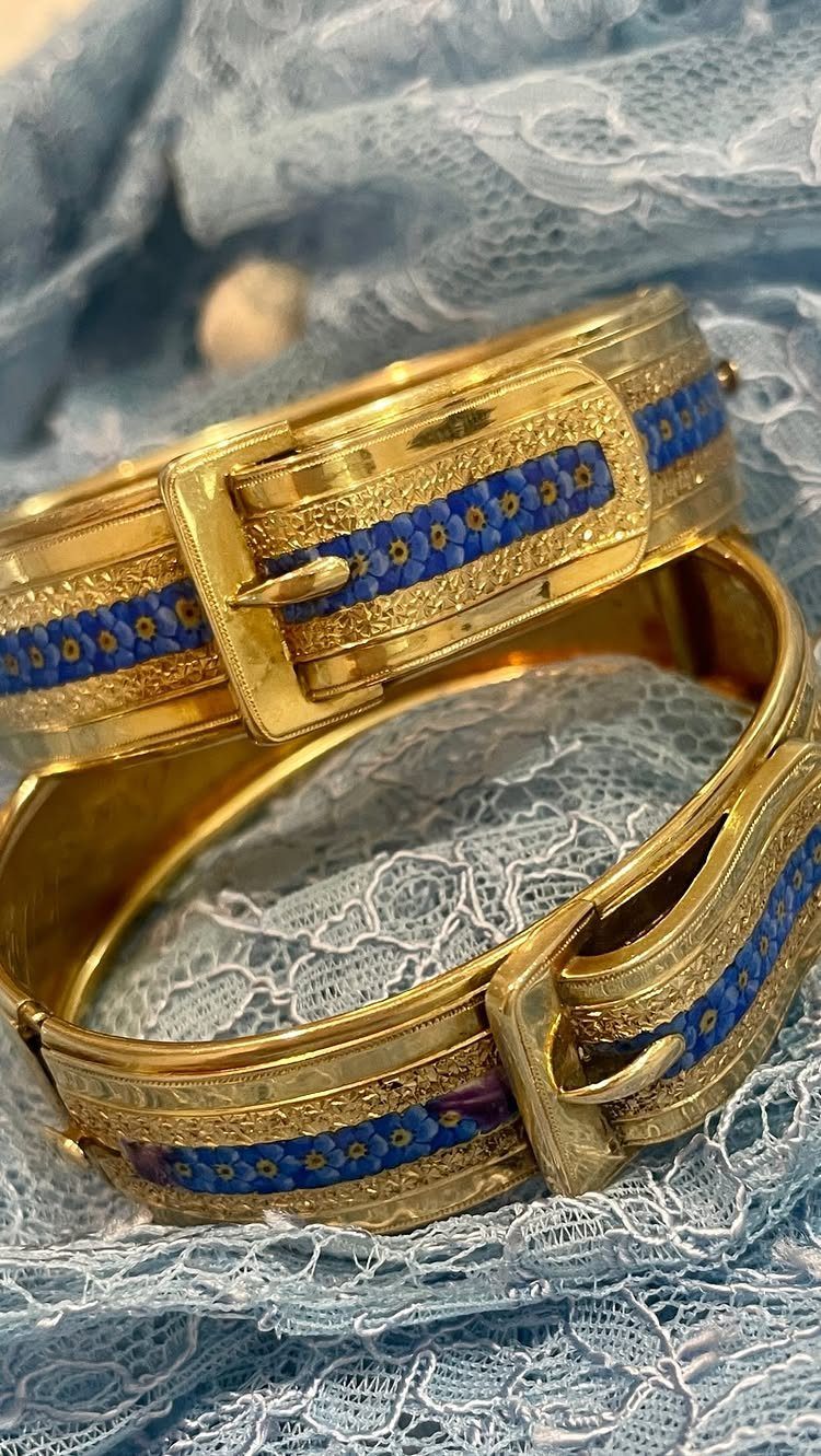 Two gold bracelets with blue floral enamel details on light blue lace fabric.