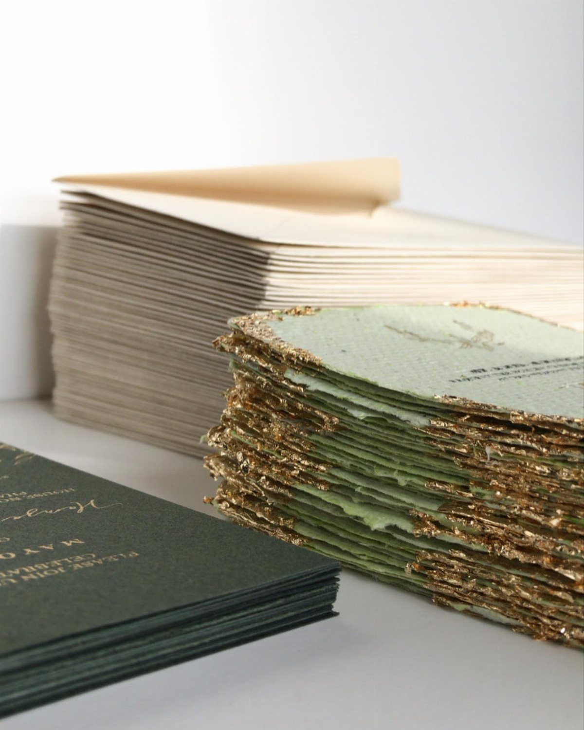 Stacks of handmade wedding invitations with gold glitter edges and sage green paper.