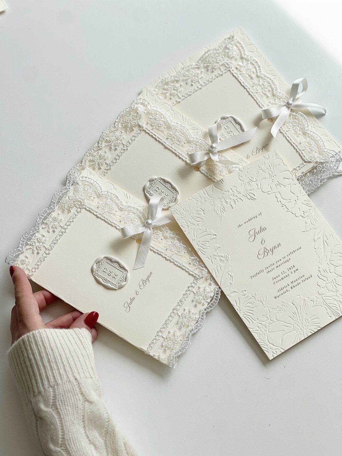 Cream wedding invitations featuring white lace trim, small bows, and gold calligraphy.
