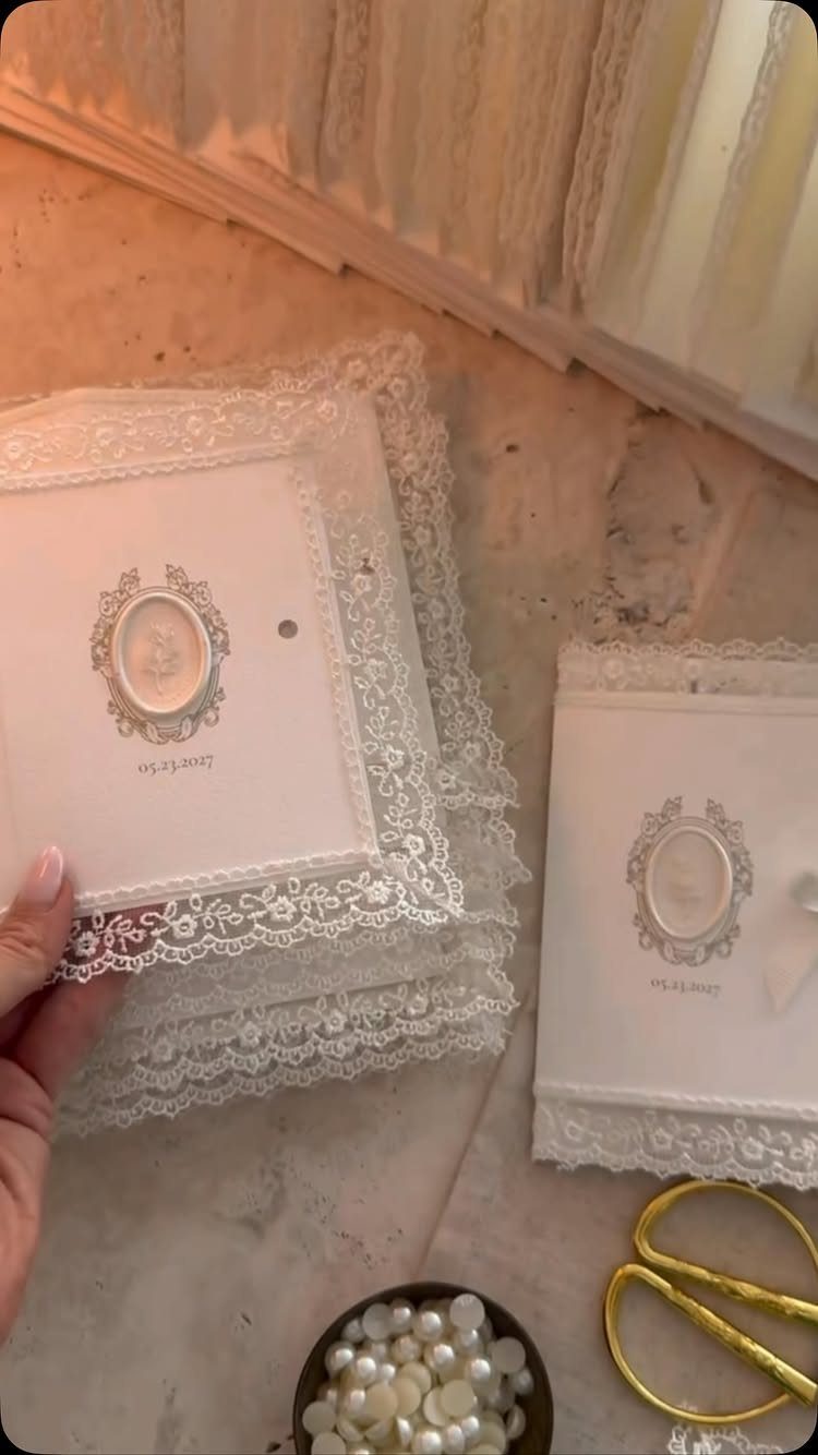 White wedding invitations with lace borders, oval portraits, gold scissors, and white pearls.