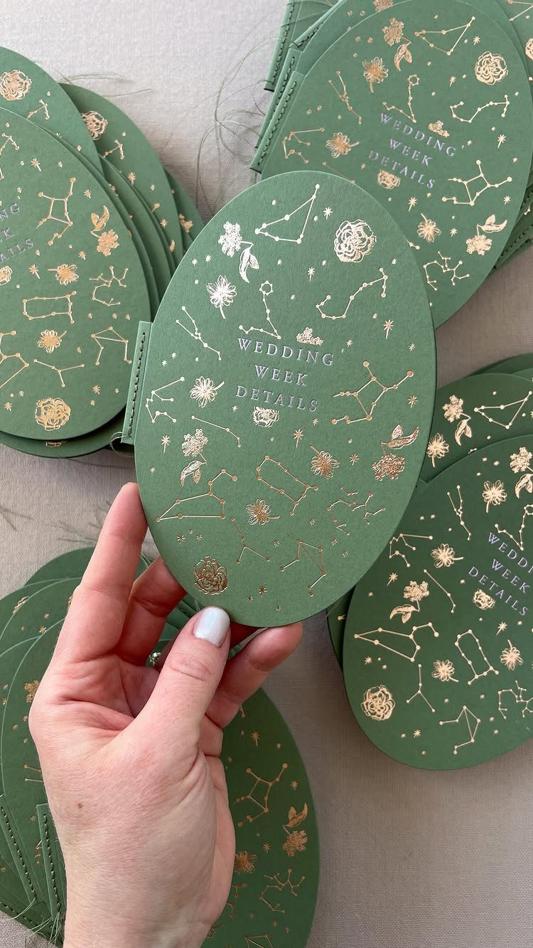 Sage green oval wedding cards with gold foil constellations and wedding week details text.