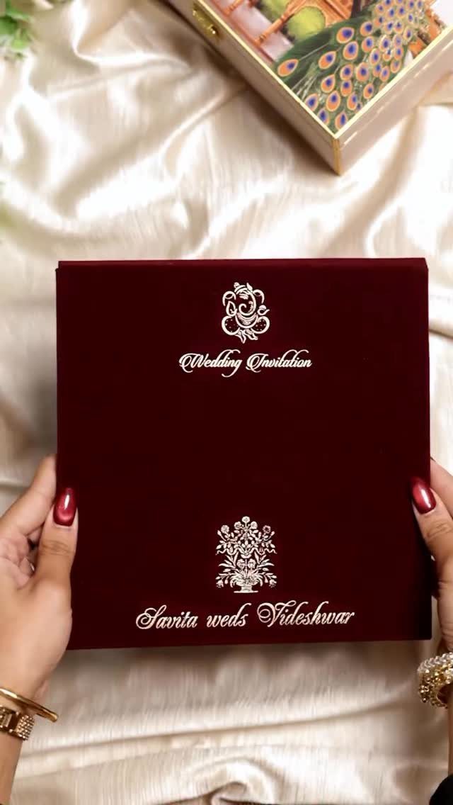 Deep red velvet wedding invitation with white ornate text held by two hands.