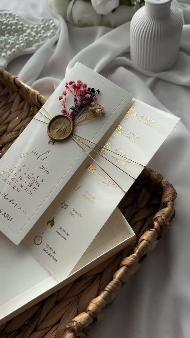 White wedding invitation cards with gold accents, wax seal, and dried pink flowers.