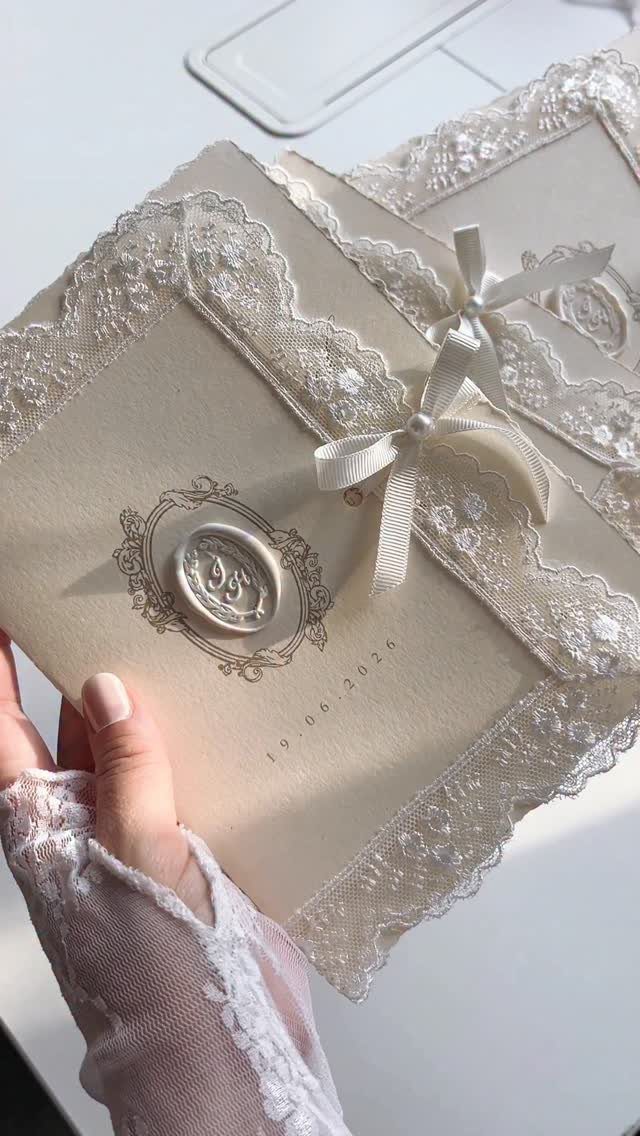 Cream wedding invitation with lace trim, satin bow, and a wax seal.