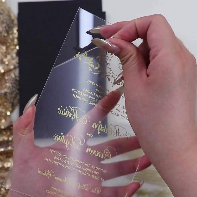 Transparent acrylic wedding invitation with gold lettering held by manicured hands.
