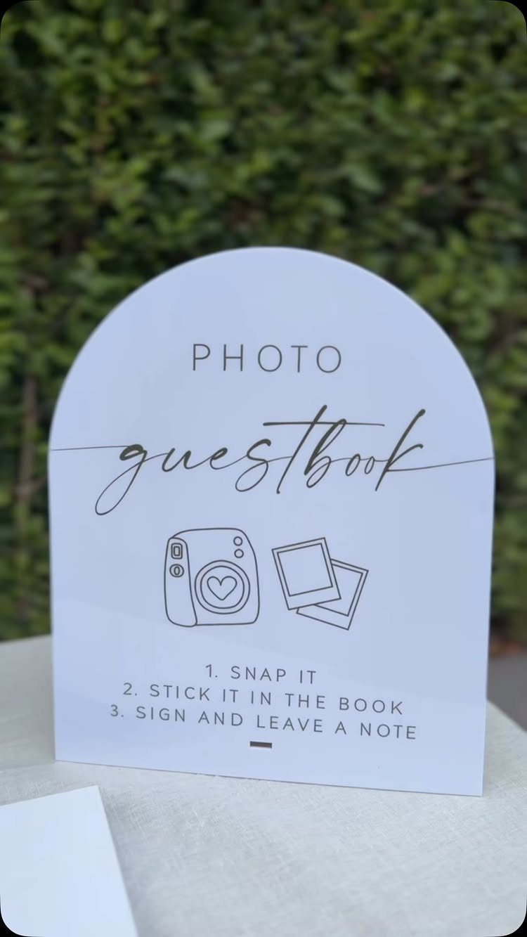 Arched white photo guestbook sign with camera illustrations against a green hedge.