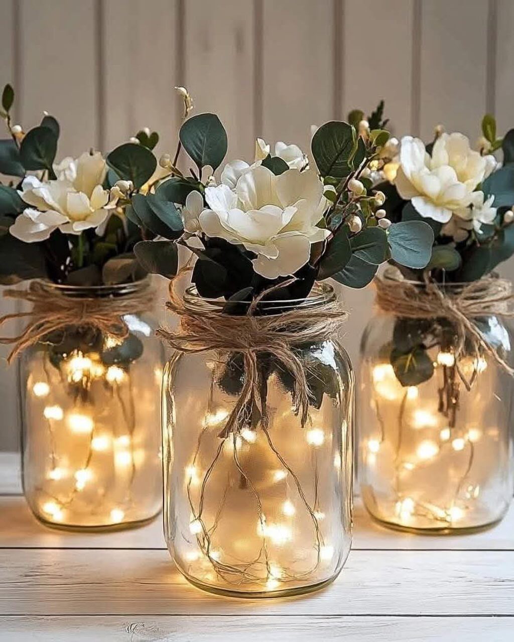 Mason jars with white flowers and fairy lights tied with twine on wood.