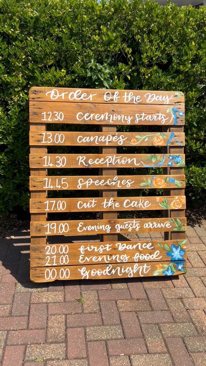 Wooden pallet sign with wedding schedule and floral paintings against a green hedge.