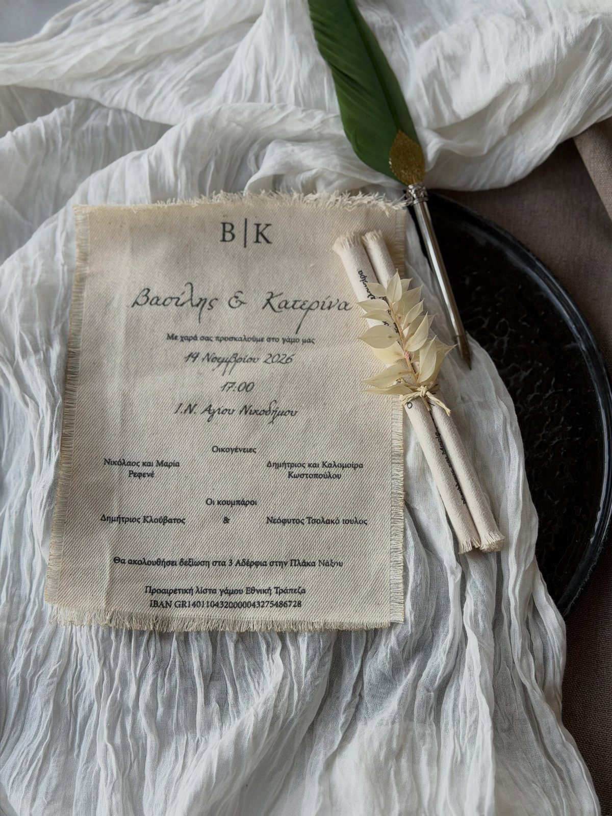 Linen fabric wedding invitation with Greek text next to rolled scrolls and quill.
