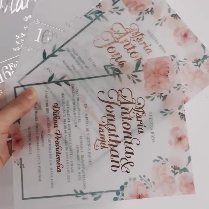 Translucent acrylic wedding invitations with gold cursive text and pink floral designs.