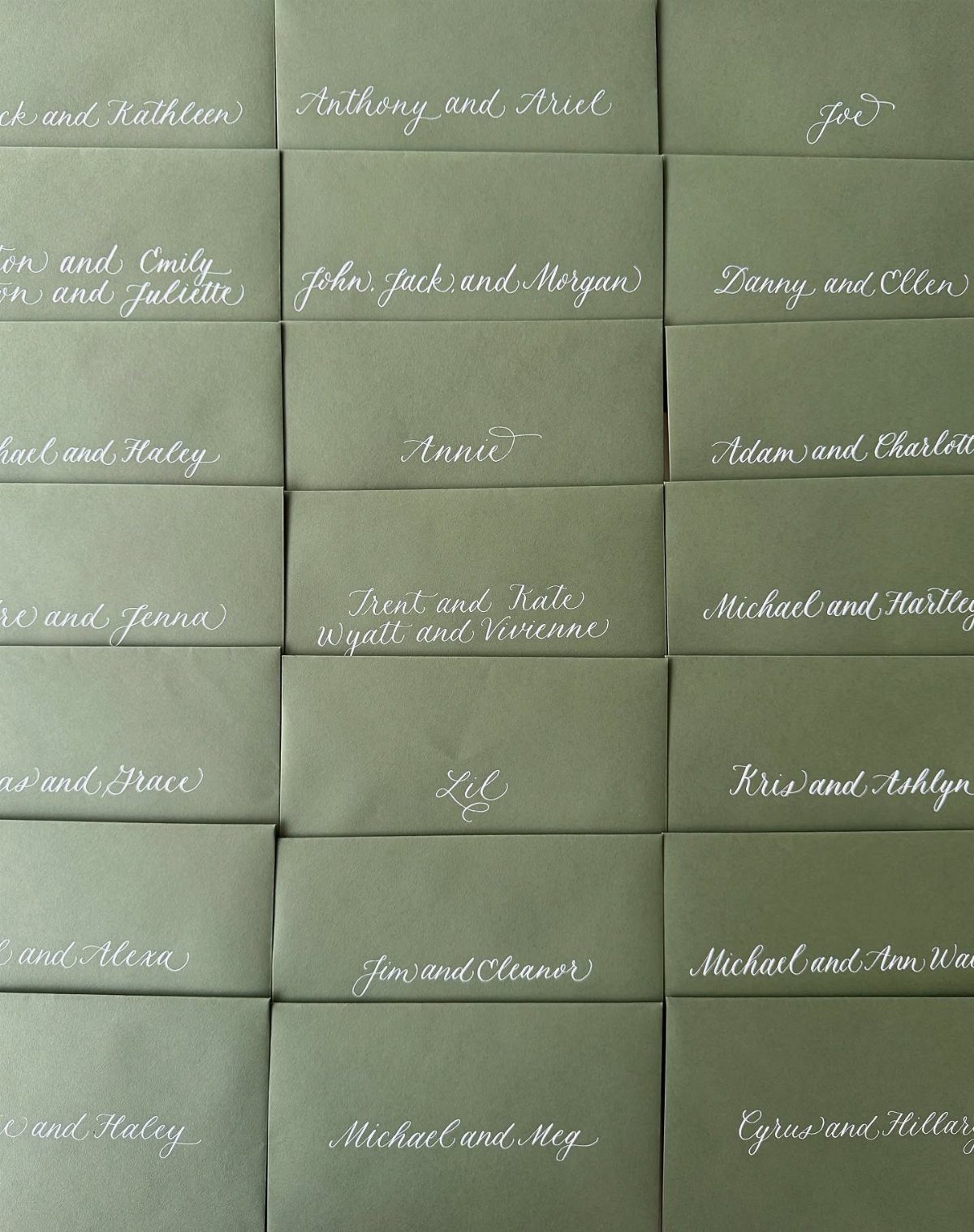 Sage green envelopes with white cursive calligraphy addressing guests for a wedding.