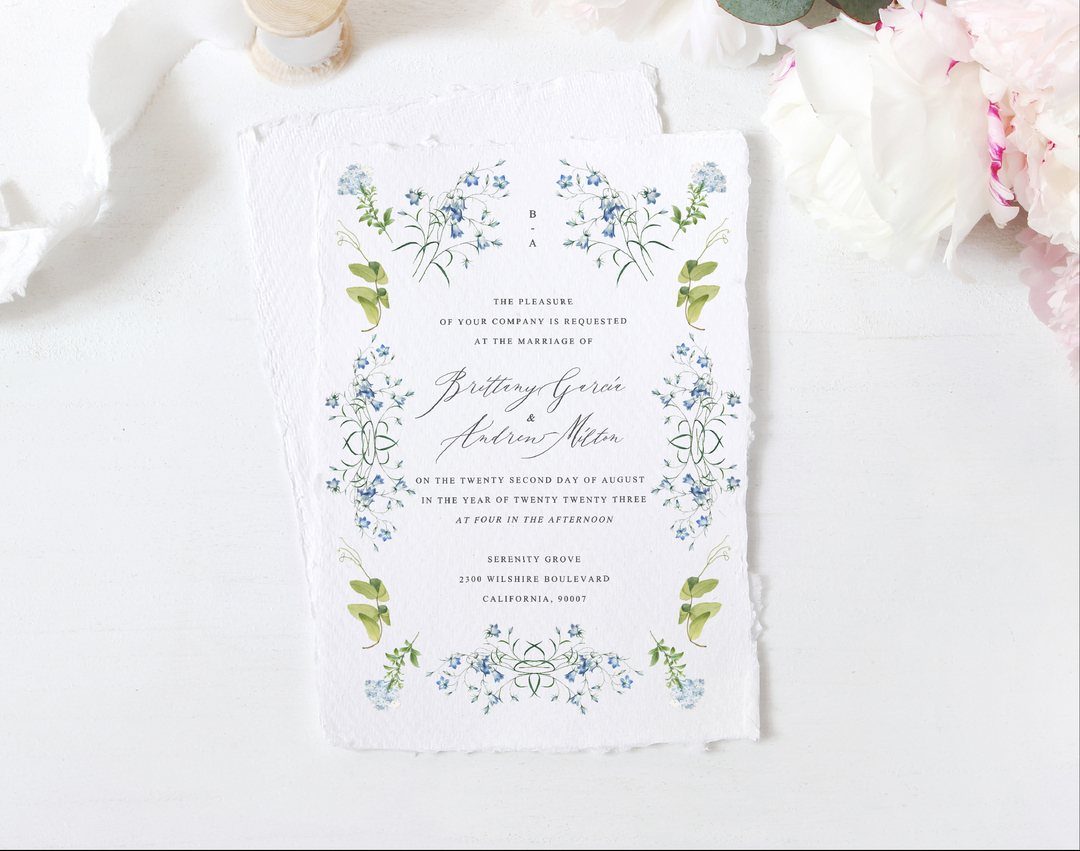 Wedding invitation with a blue floral wreath border and deckle edges near peonies.