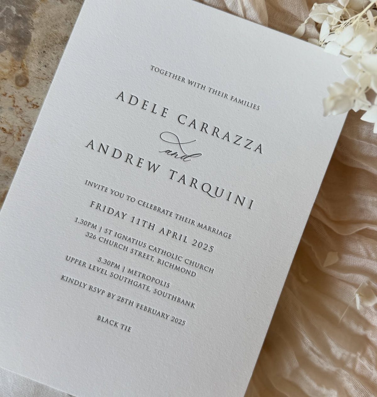 White wedding invitation card with black text resting on beige fabric and flowers