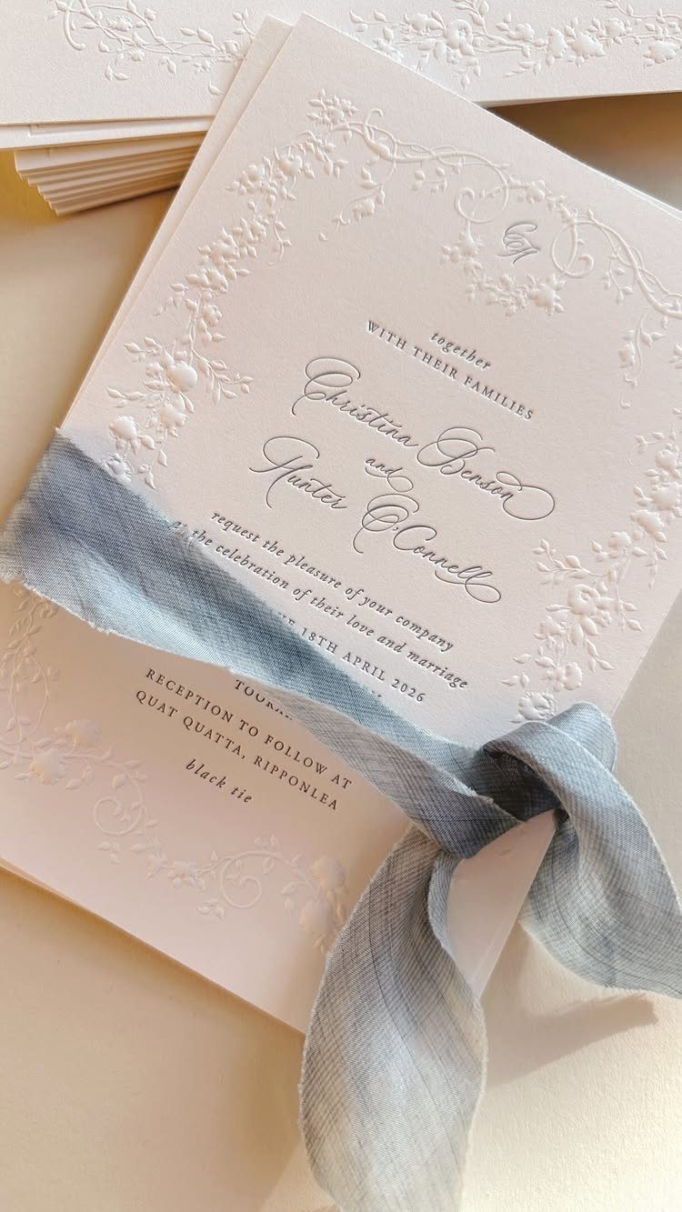 White embossed wedding invitation tied with a dusty blue silk ribbon on beige background