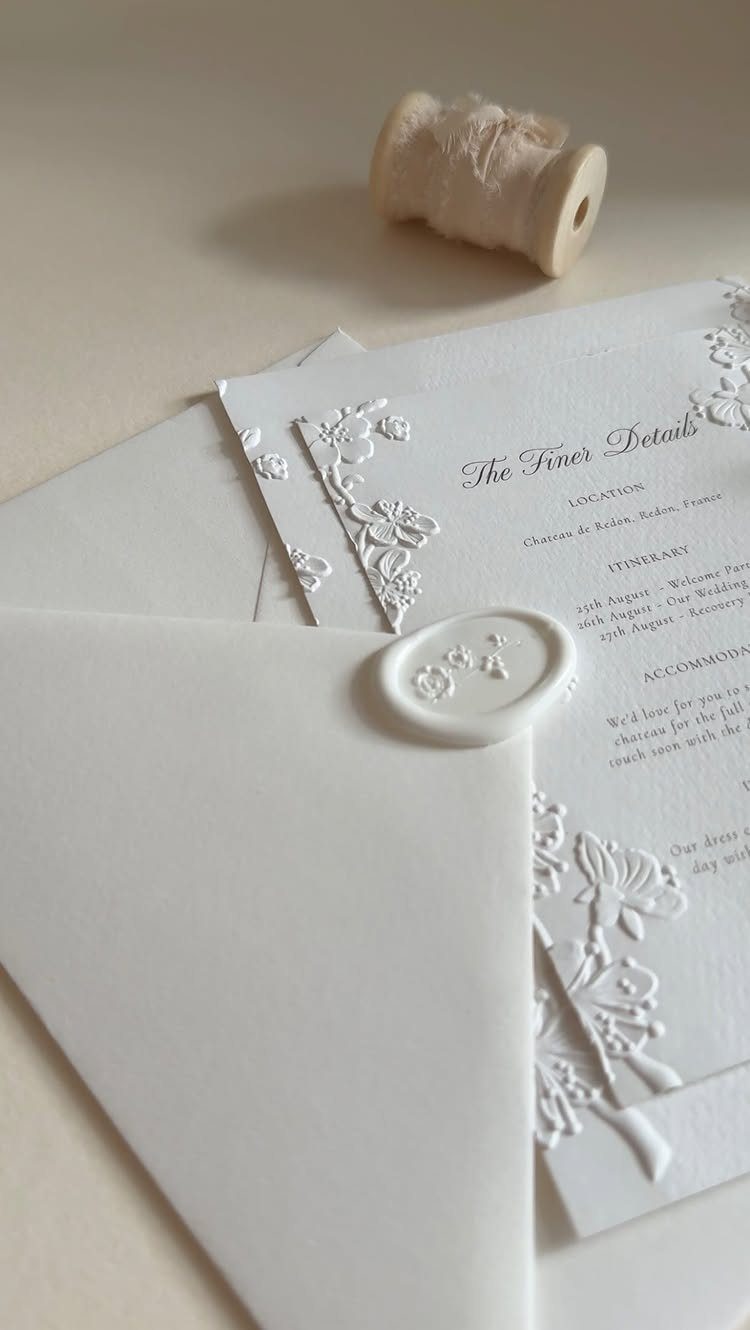 White wedding invitations with embossed floral details, a wax seal, and white twine.