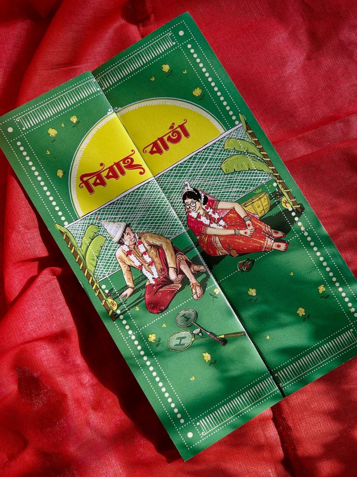 Green Bengali wedding invitation card with traditional illustrations on red fabric background