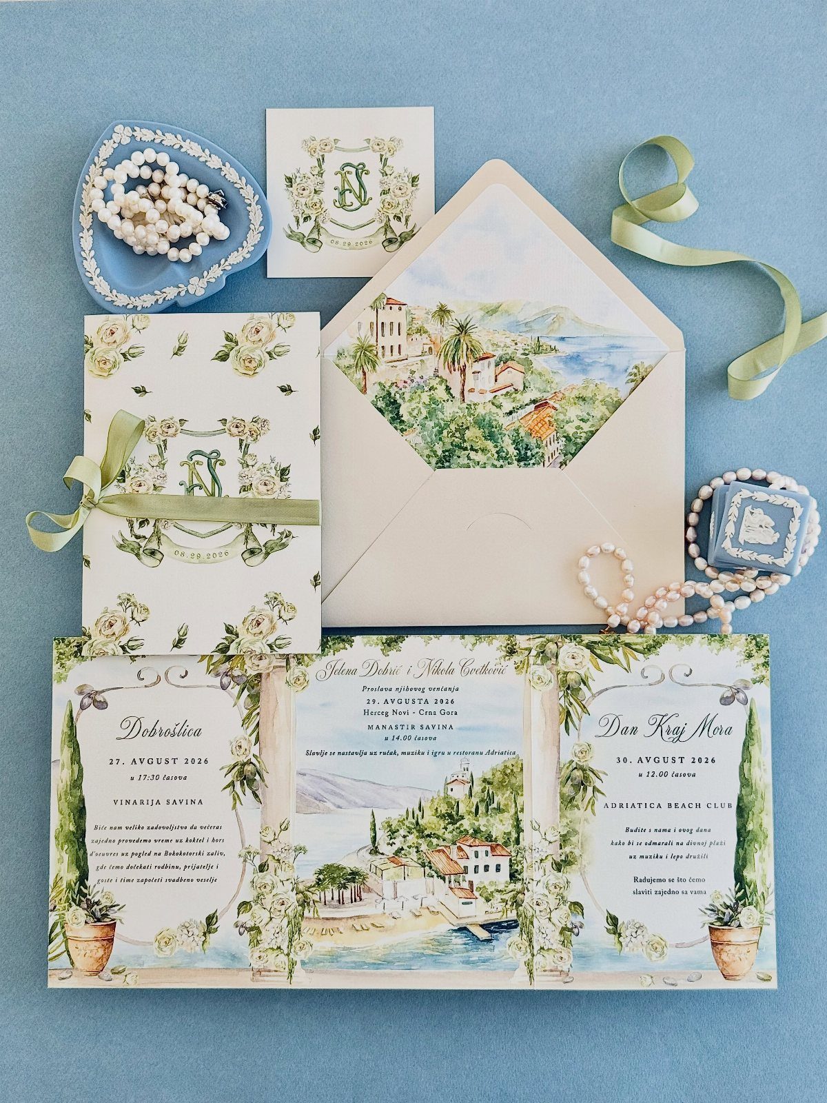 Wedding invitation suite with coastal village illustrations, green ribbons, and pearl accessories.