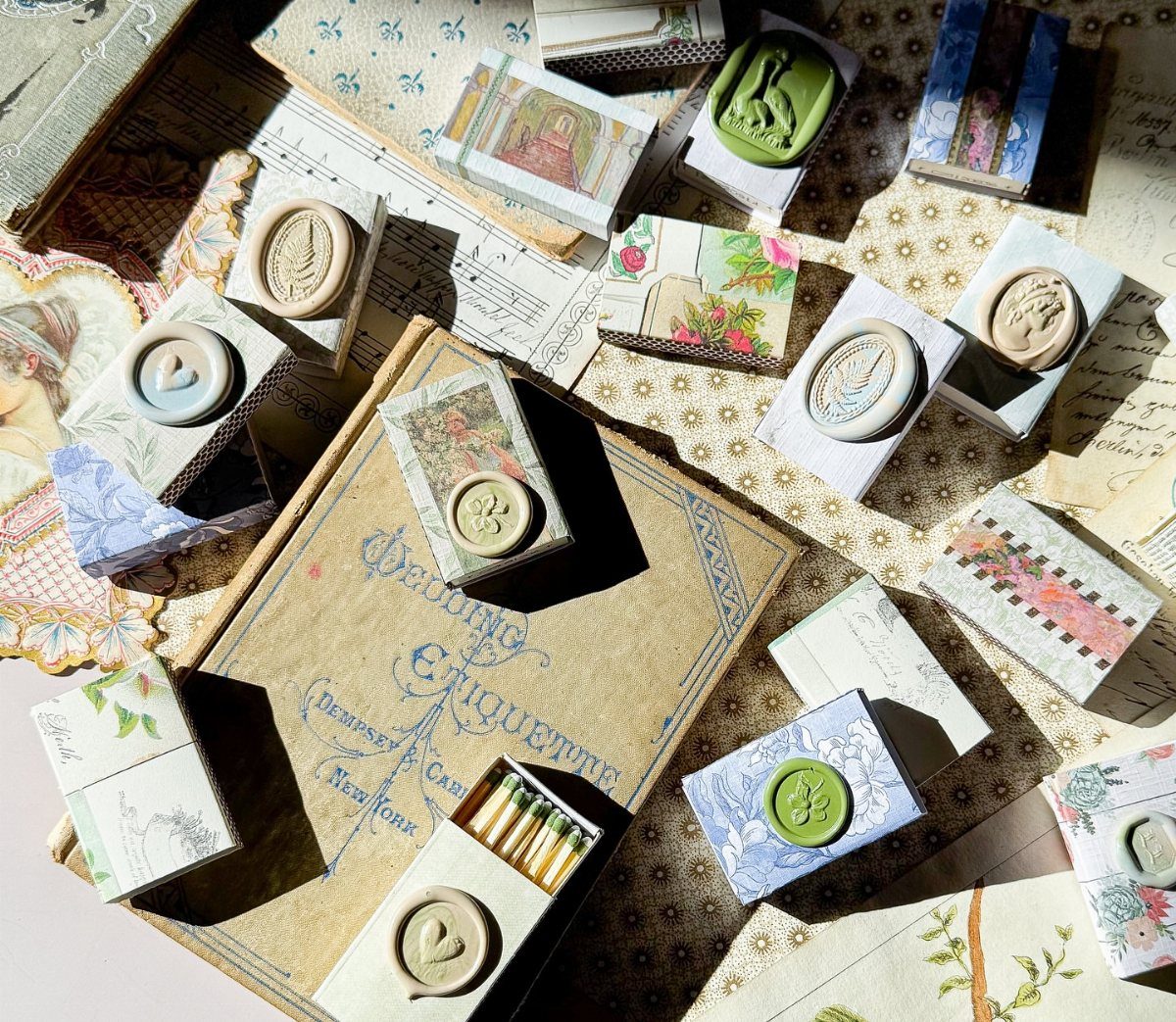 Decorated matchboxes with wax seals scattered on vintage books and patterned paper.