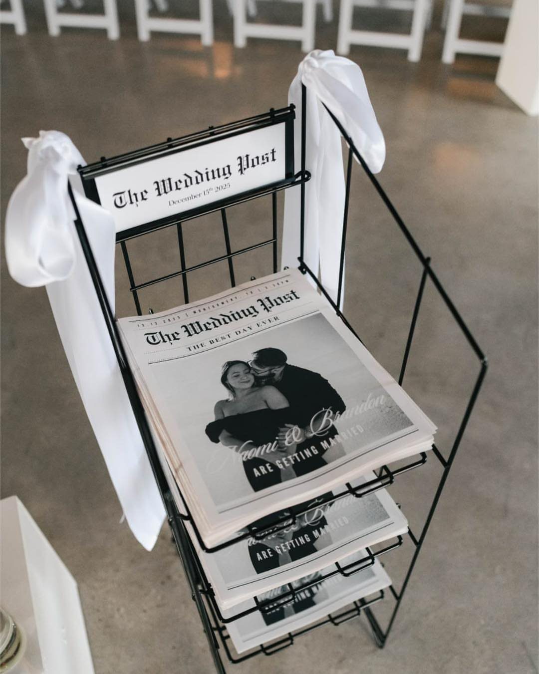 Wedding newspapers in a black wire rack decorated with white ribbons.