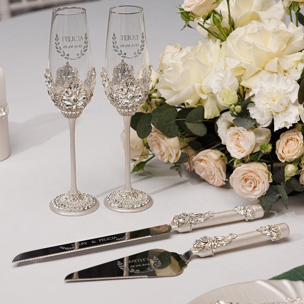 Crystal embellished champagne flutes and cake serving set next to white roses.