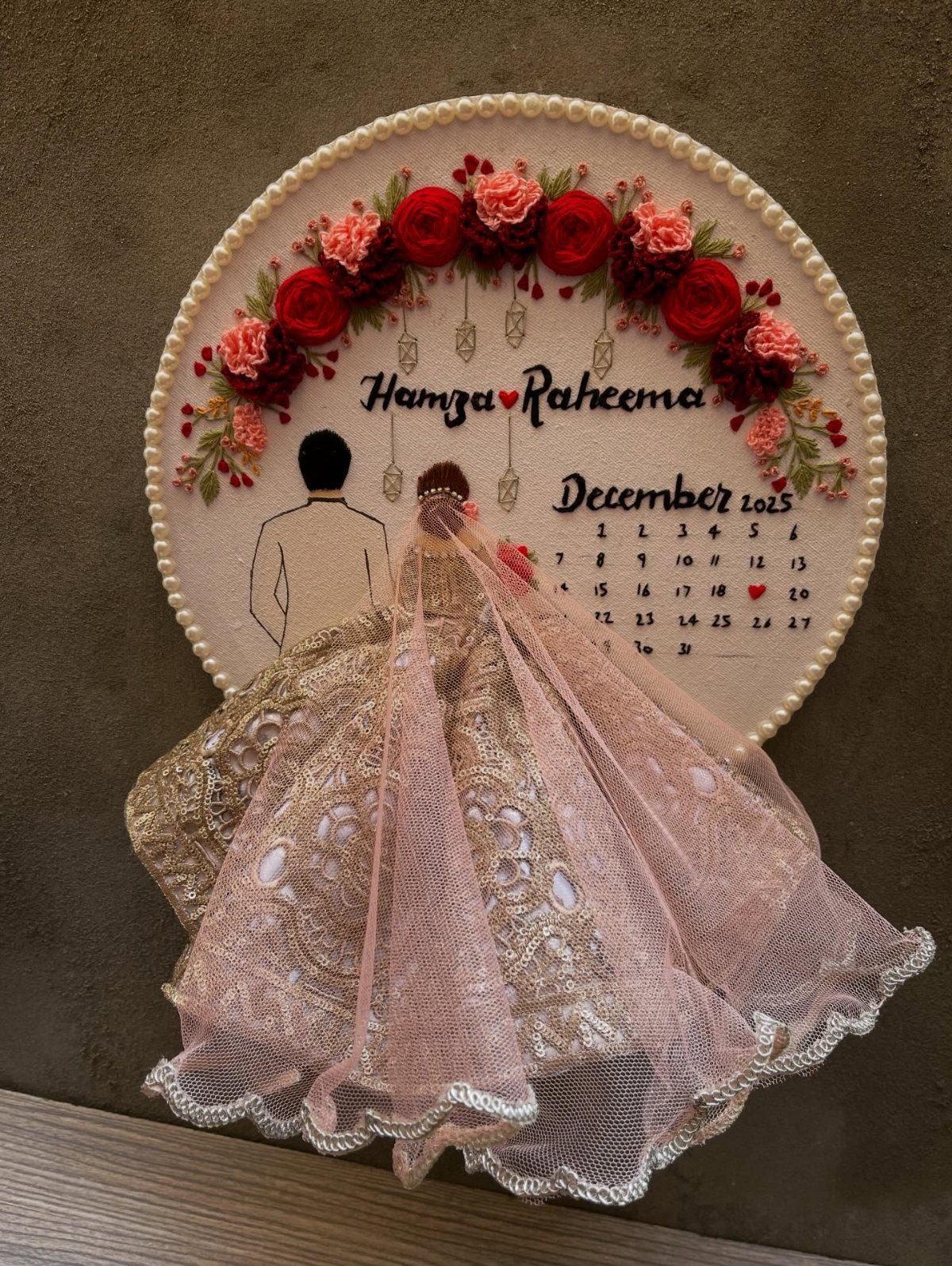 Embroidered circular wedding art with red roses, names, calendar, and lace veil.