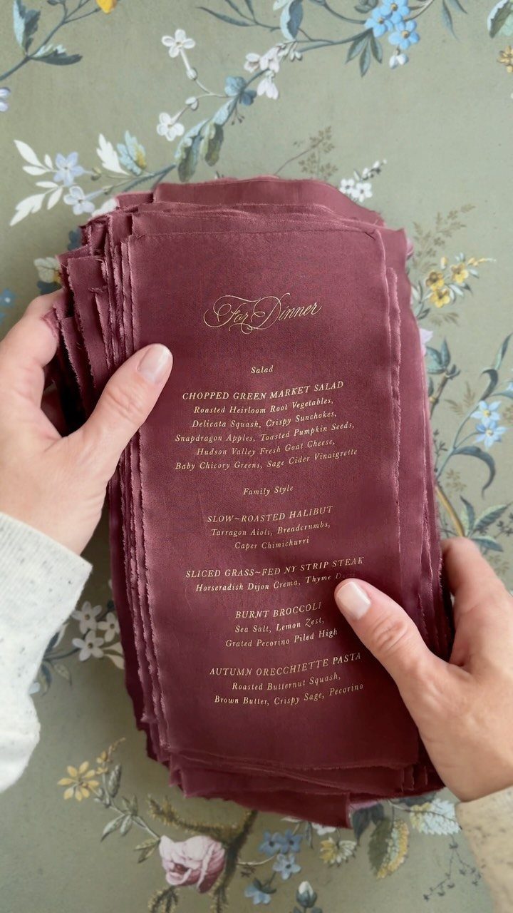 Burgundy paper menus with gold foil text and deckled edges held by hands.