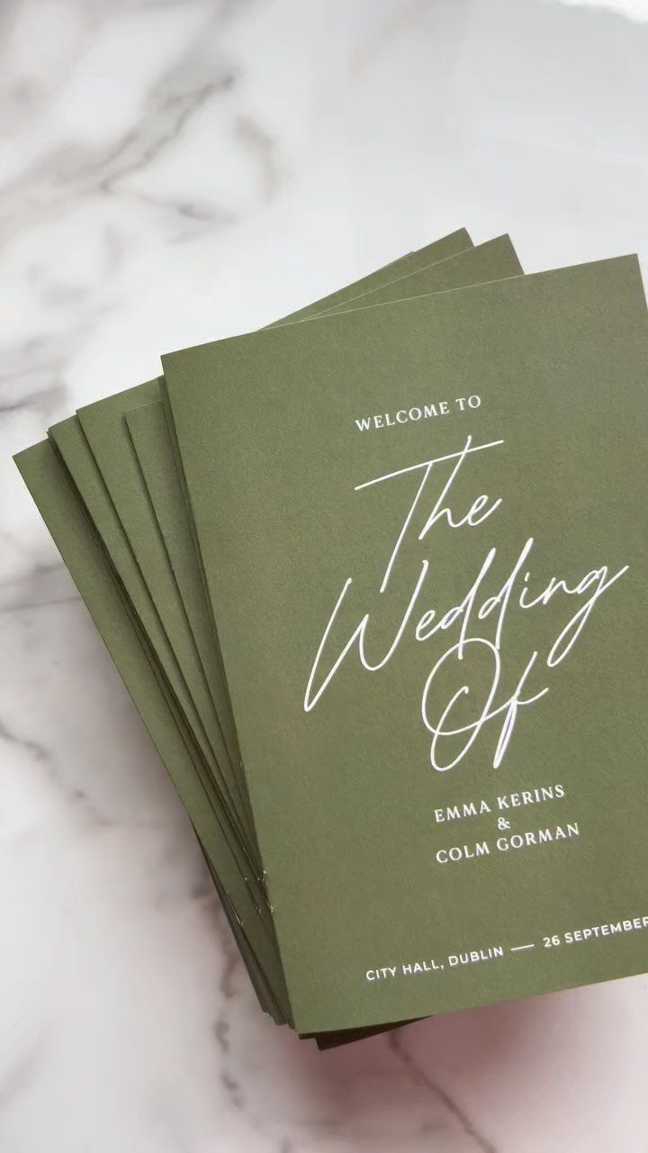 Olive green wedding programs with white script text on a white marble background