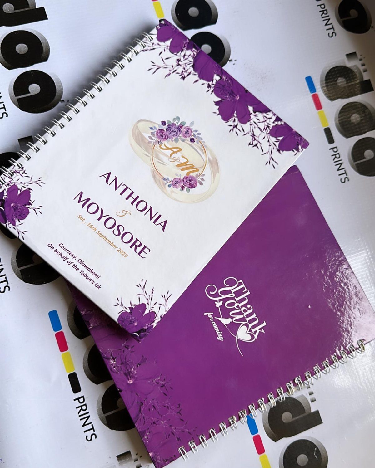 Spiral-bound wedding programs featuring purple floral designs and a thank you message.