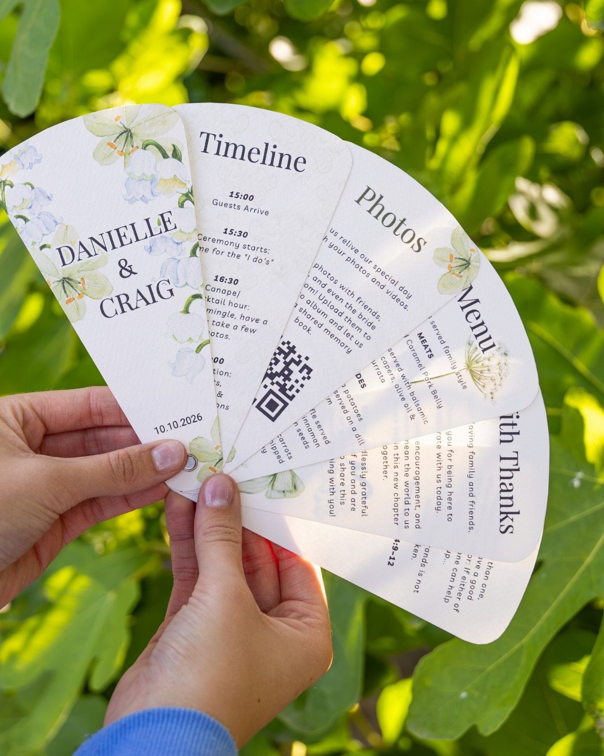 Cardstock wedding program fan with floral accents and event timeline held against greenery.